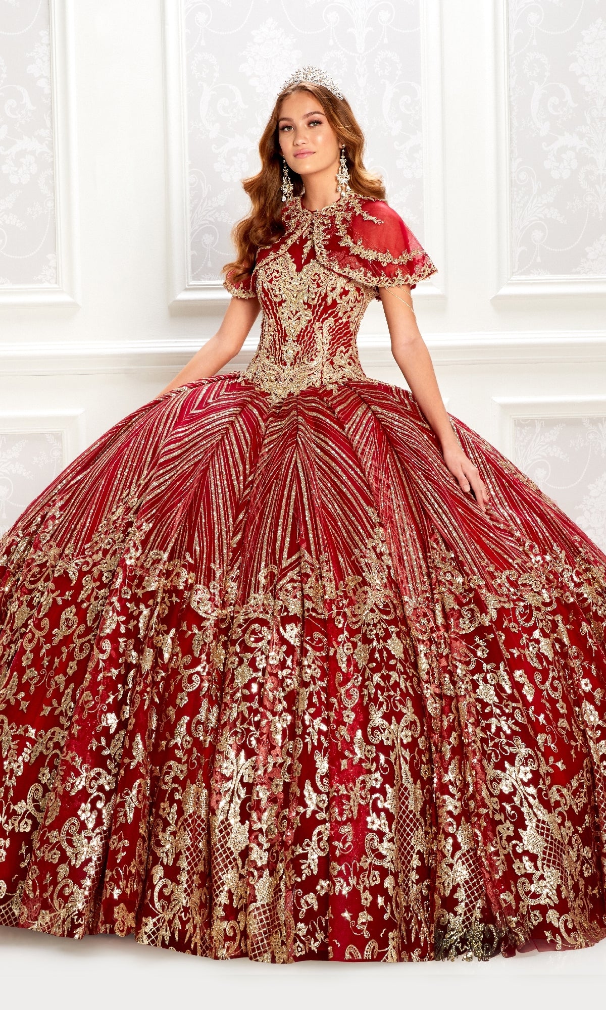A woman models the Princesa PR22034, a wine red quinceanera dress with gold embellishments, full skirt, and a removable bolero jacket, standing indoors against an ornate white wall.